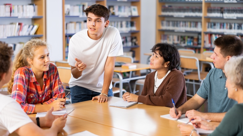 Positive teenage male student giving report during group lesson in college library